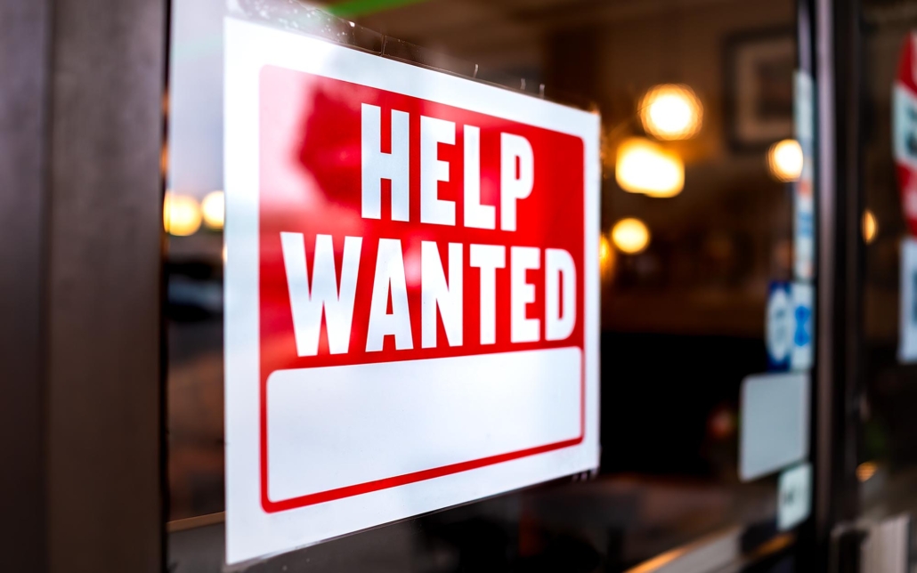 Image of a help wanted sign posted on the front of a retail store.