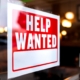 Image of a help wanted sign posted on the front of a retail store.