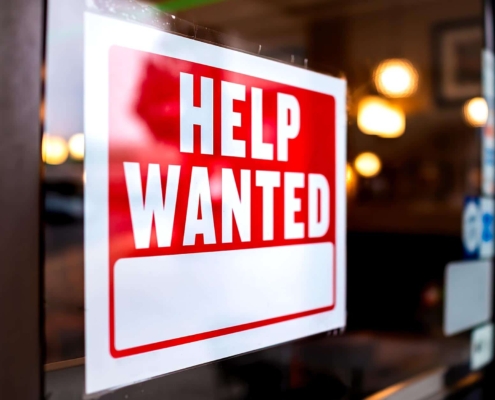 Image of a help wanted sign posted on the front of a retail store.